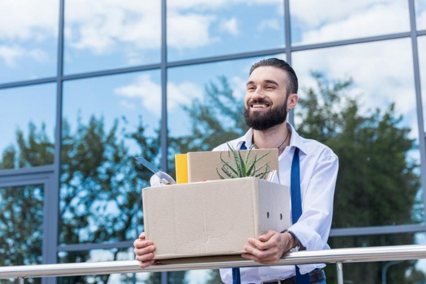 Man smiling, holding a box of office supplies that would belong on a desk at work.